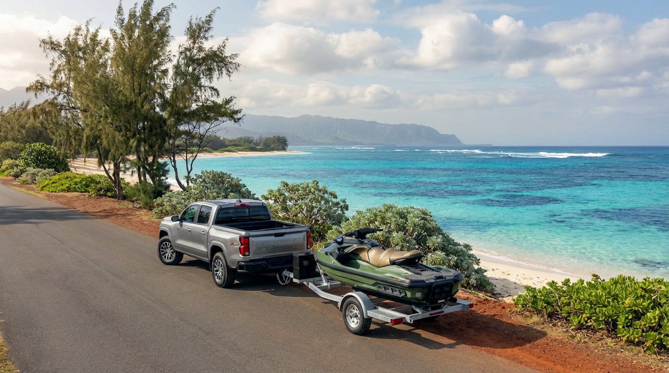 A silver pickup truck towing a green jet ski on a trailer on a coastal road overlooking a turquoise ocean.