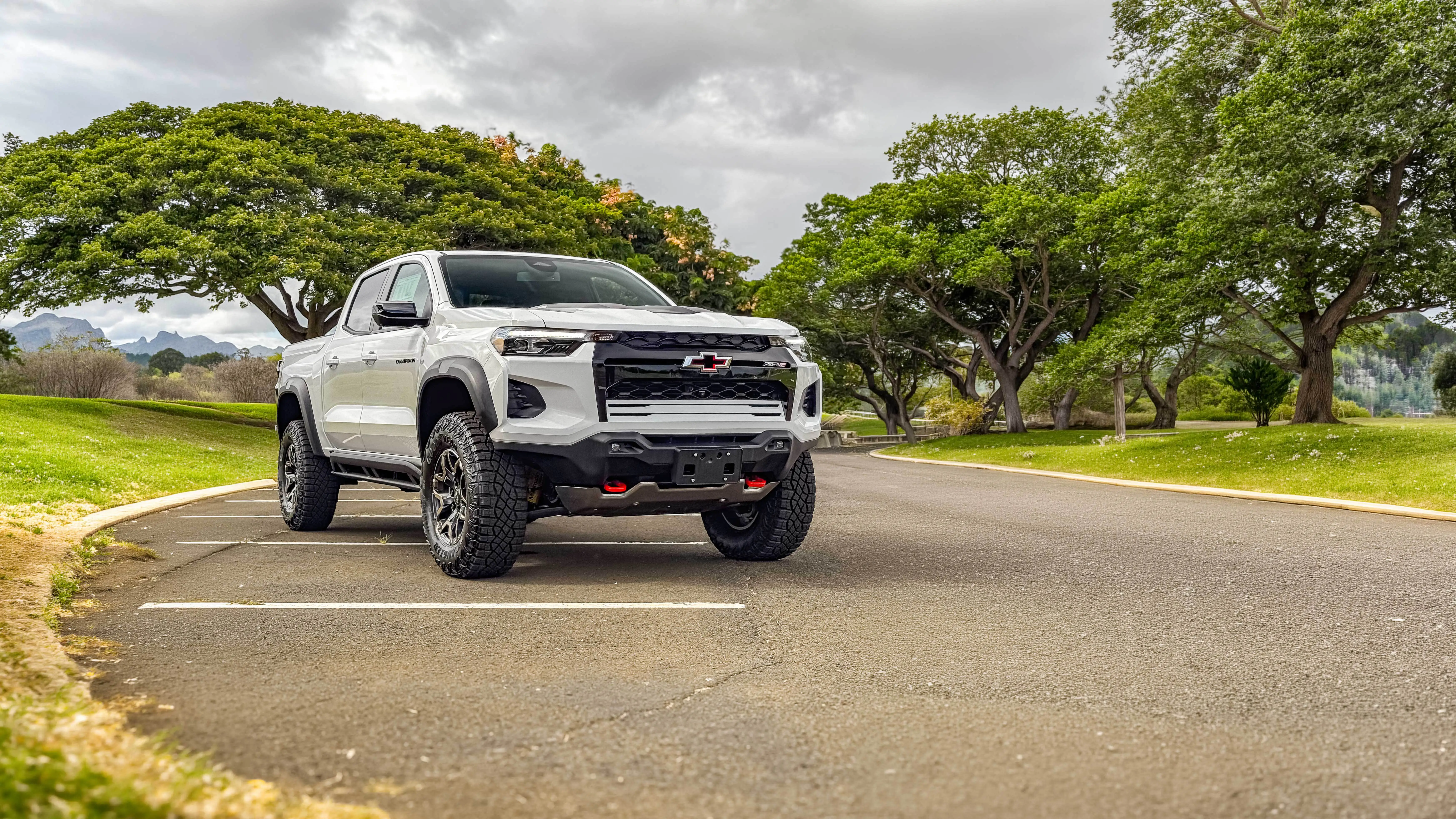 White Chevrolet Colorado pickup truck parked on asphalt with large green trees, grass, and distant mountains.