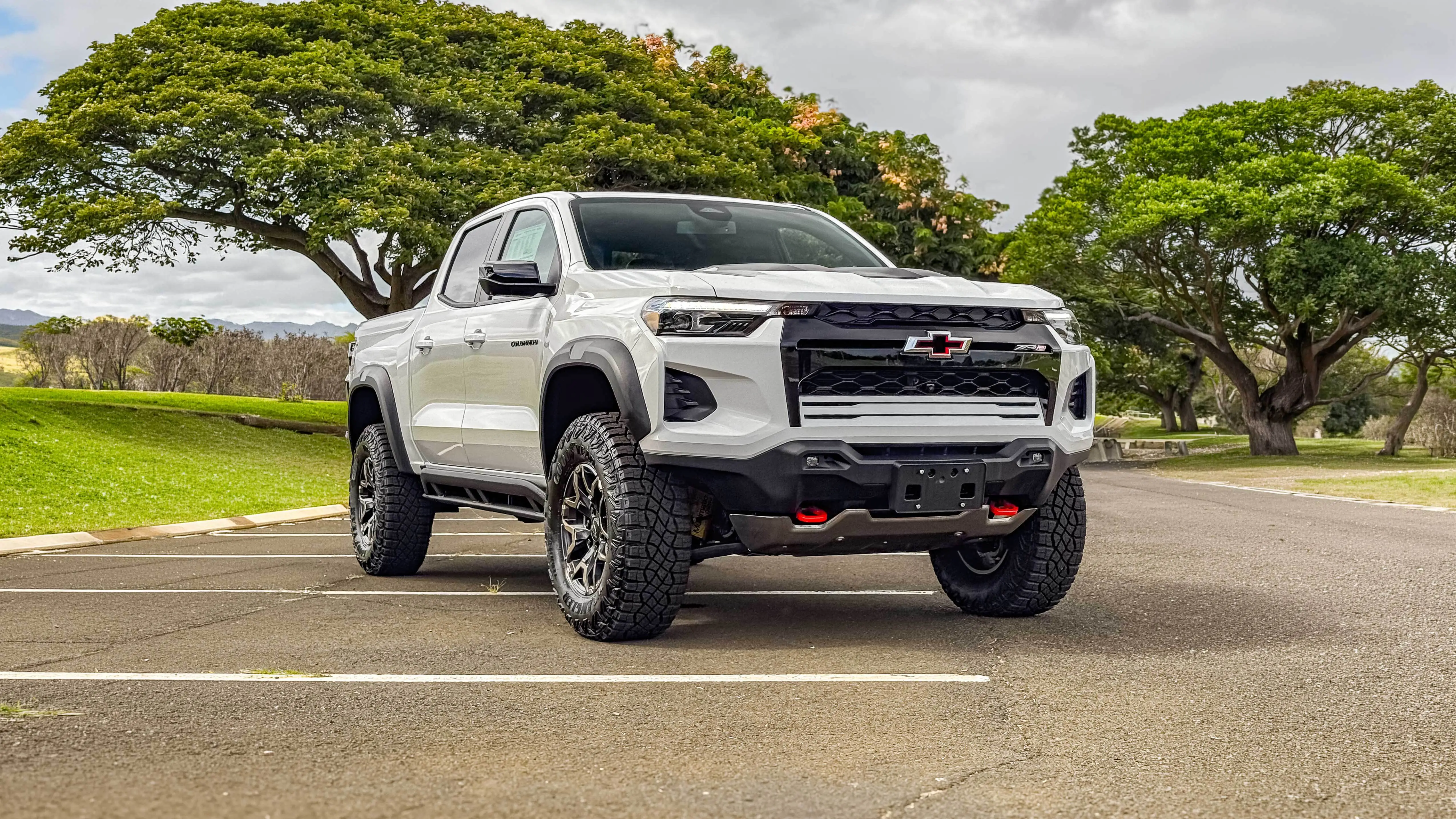 A white Chevy Colorado pickup truck parked in a lot, with large trees in the background under a cloudy sky.